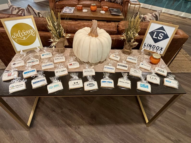 Table with logo'd cookies on top. In center is white pumpkin. To left is sign that says 'welcome' and to right is sign with Lindox Siegel logo.