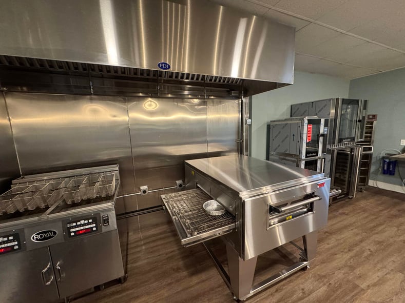 Range and conveyor oven in Baker Culinary Club. Brown wood floor is in center and other foodservice equipment is to the right of ovens.